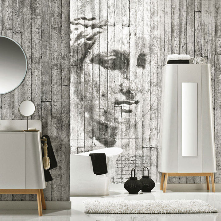 Modern bathroom interior with artistic face mural on rustic wooden plank wall, freestanding white bathtub, minimalist vanity with round mirror, tall cabinet, black towels, and stylish decor accessories.