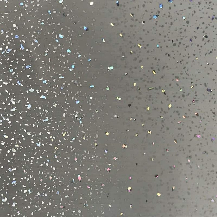 Close-up of a gray surface speckled with colorful glitter, featuring a variety of sparkly specks and holographic confetti scattered across a neutral background.